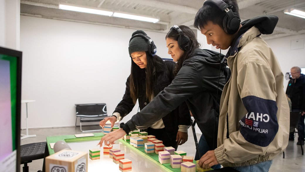 Three students wearing headphones smile as they place blocks onto a light table.