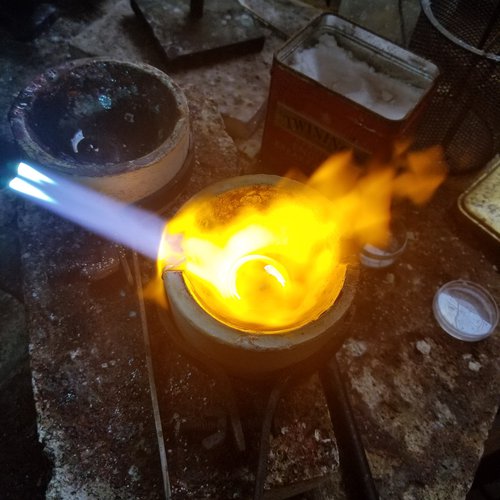 Gold being recycled by melting is on fire and illuminated by the blue light of a melting torch in Curtis Arima’s studio.