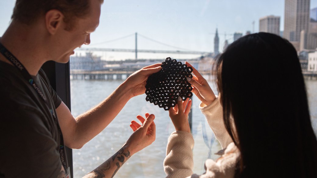 Two students hold up a 3-d protoype of a hexagonal mesh against a window with the SF Bay Bridge in the background.