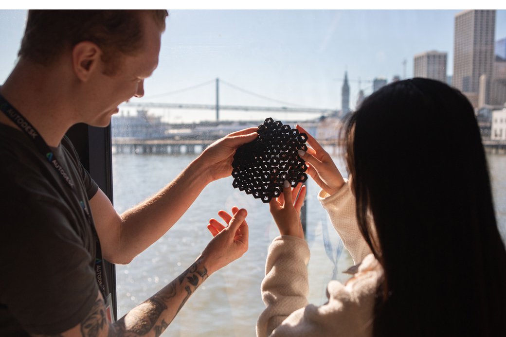 Two students hold up a 3-d prototype of a hexagonal mesh against a window with the SF Bay Bridge in the background.
