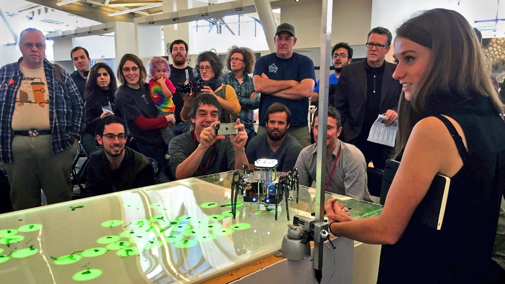 A crowd watches as a small robot walks on a screen displaying small green dots.