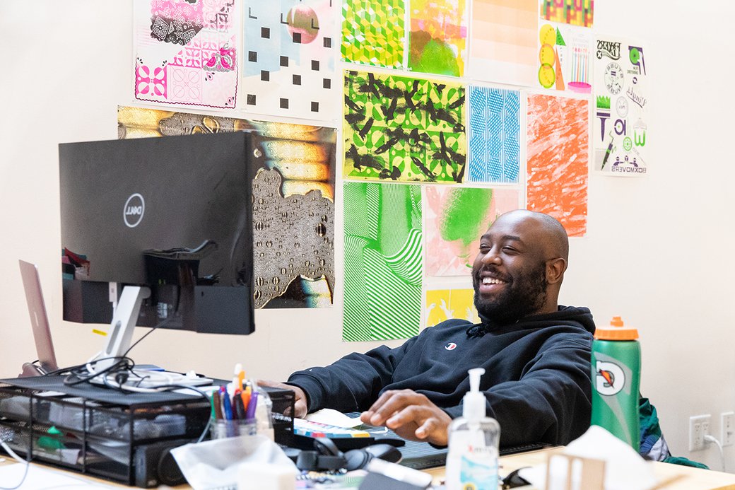 Design student smiling at his computer desk. The wall behind him is full of colorful posters.