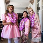 Three students pose in pink silk dresses and flower leis.