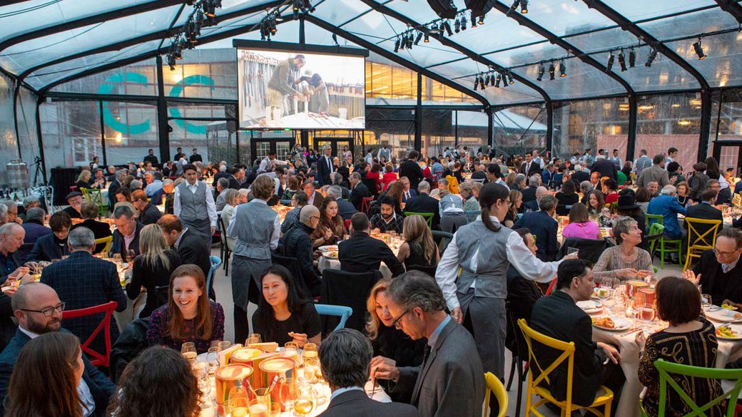 wide shot of a room full of people eating as a video plays on an elevated projector in the background