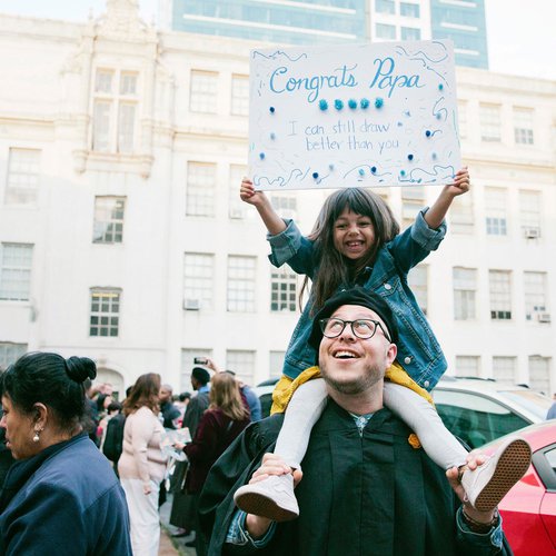 A child sits on her graduating father's shoulders while holding a silly sign congratulating him.