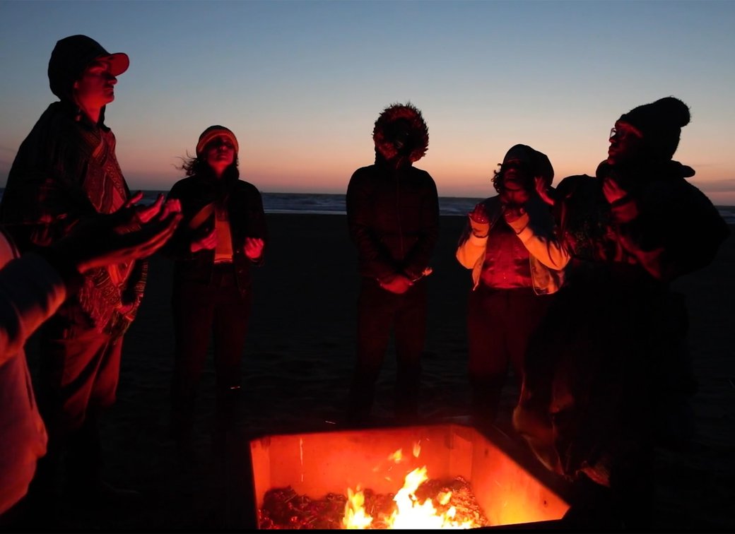 A group of people perform a fire ceremony on Ocean Beach at dusk.