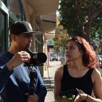 Two people, Jeremiah Ysip and Kat Cole, stand on a sidewalk discussing a scene happening out of view. Jeremiah is holding a camera.