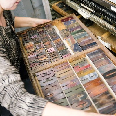 A person holds a large flat box of ink-covered wooden letters.