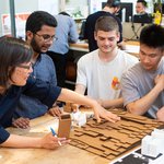 Students gathered around a small, wooden architecture project in a classroom.