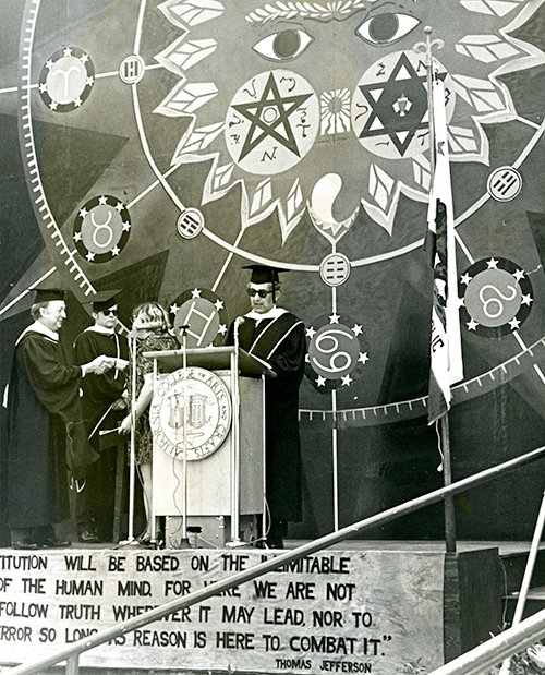A person in a cap, gown, and sunglasses stands giving a speech at a lectern in front of a mural of a sun.