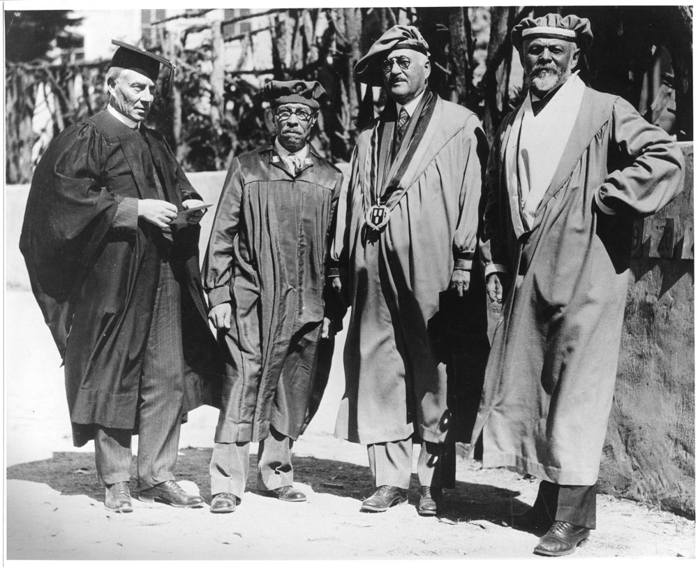 Four men in commencement gowns and tams stand in a line and pose solemnly in a black and white aged photograph.