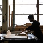 A student cuts wood wearing goggles and a face mask in a light-filled workshop.