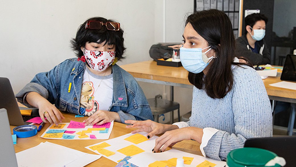 Two students sit at a workbench, glancing between a computer and post-its arranged neatly on paper.