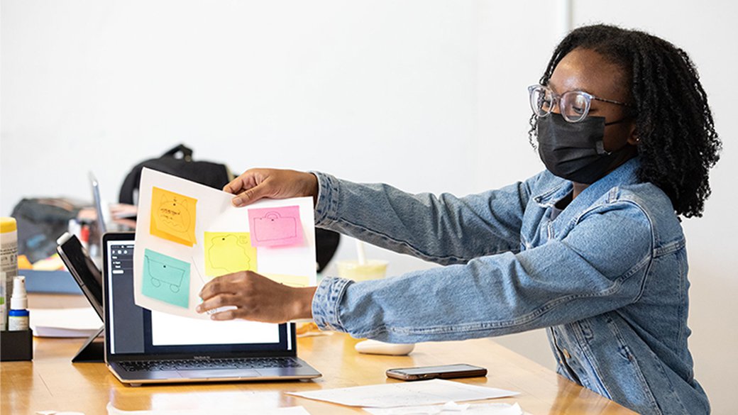 A student wearing a denim jacket holds up a piece of paper with post-its of drawings neatly arranged.