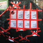 Photograph of a fence covered in black tarp overlaid by red tape placed in X shapes and a grid of posters.