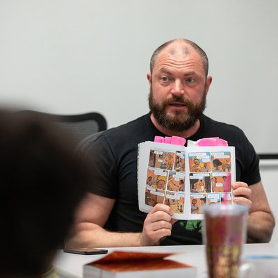 Justin Hall—a man with a beard, wearing a black t-shirt—holds up a graphic novel while speaking to a class.