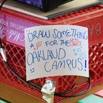 A photo of a hand-drawn sign on a red carton that says, “Draw something for the Oakland campus!”