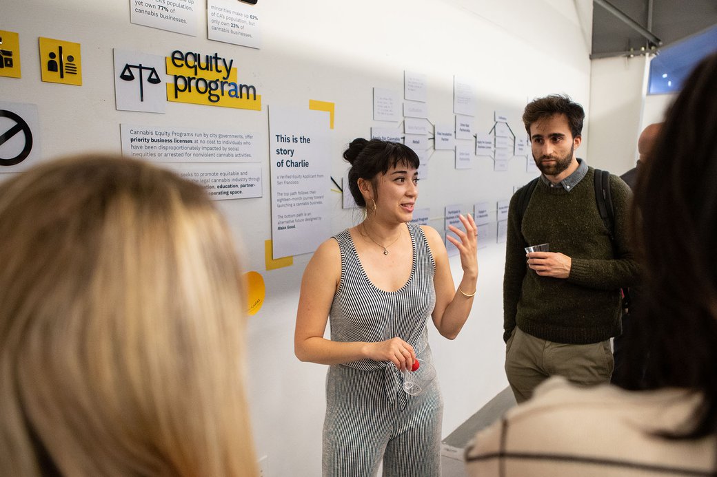 A woman with dark hair and a black-and-white striped jumpsuit talks about her project to a small group of people.