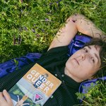 The author lays in the grass holding their book (Gender Queer) with the cover facing the camera.