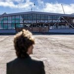 Architect Jeanne Gang stands with her back to the camera as she visits the construction site of the new “Double Ground” expansion project for California College of the Arts in San Francisco. She looks toward the current building across a flat, dirt lot where the new campus will be built.