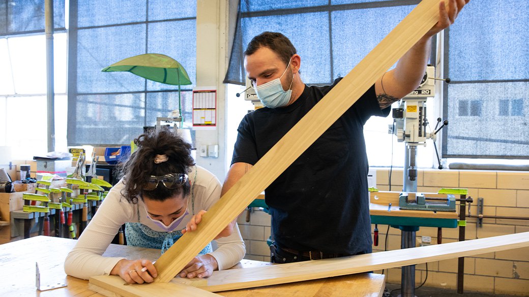 The furniture shop manager holds a long piece of wood while a student attaches it to a 2 by 4.